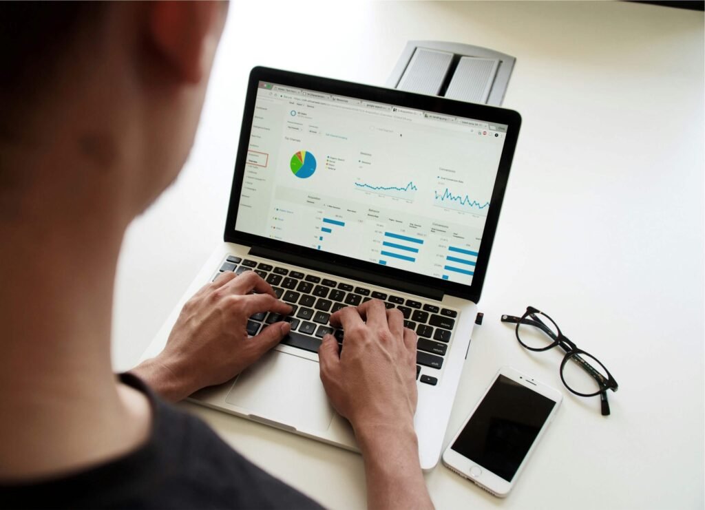 A person typing on a laptop displaying analytics dashboards and charts, with a smartphone and eyeglasses placed on a white desk nearby
