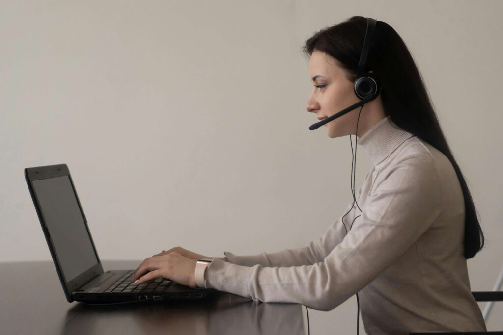 A woman wearing a headset works on a laptop at a desk, appearing to assist customers or attend an online meeting in a minimal workspace